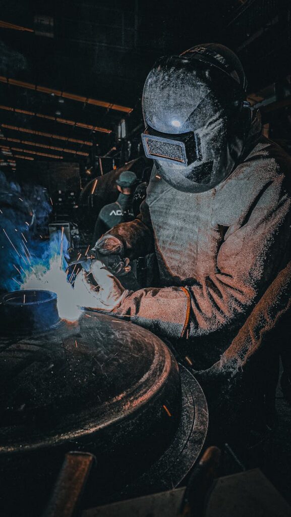 pexels-photo-31349534 A welder in protective gear working on a metal piece in a workshop with bright sparks.